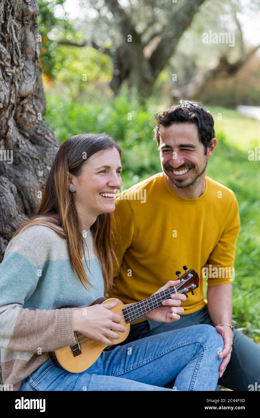 Cheerful man looking at girlfriend playing guitar while sitting in countryside Stock Photo