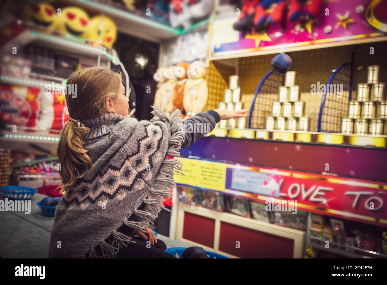 Young girl in poncho sweater at fair booth throwing ball while playing knockout game. Munich, Germany Stock Photo