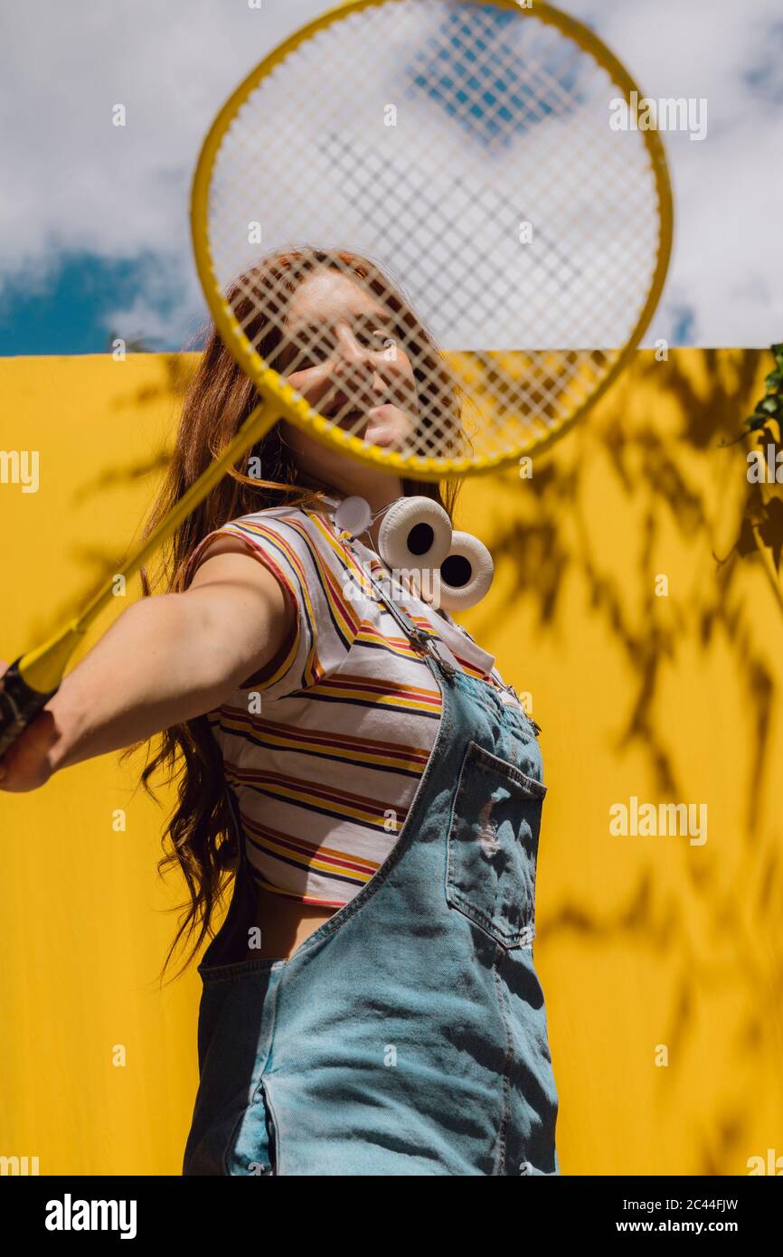 Young woman looking through badminton racket over yellow wall during sunny day Stock Photo