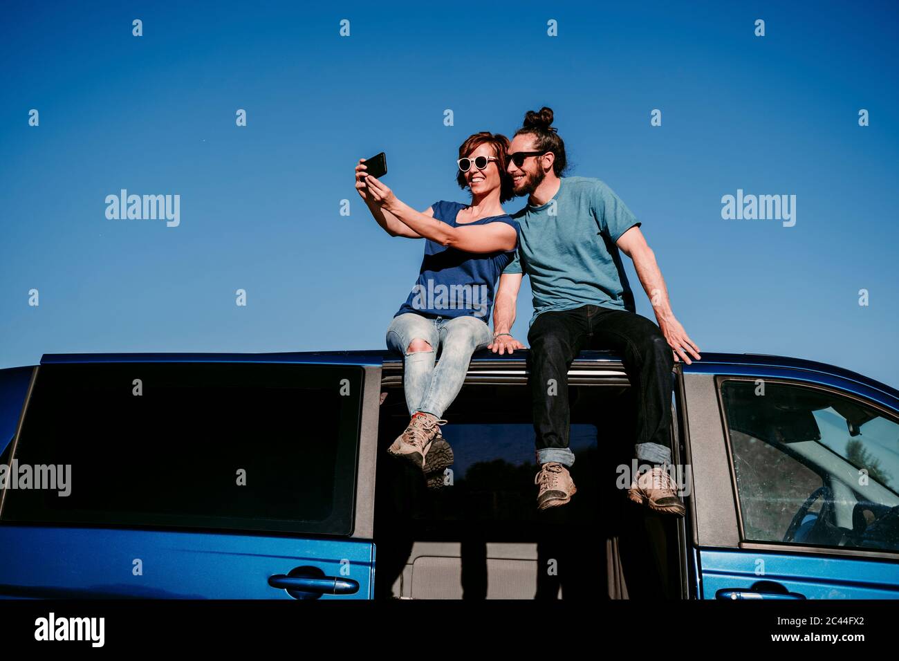 Happy couple sitting on the roof of a minivan taking a selfie Stock Photo