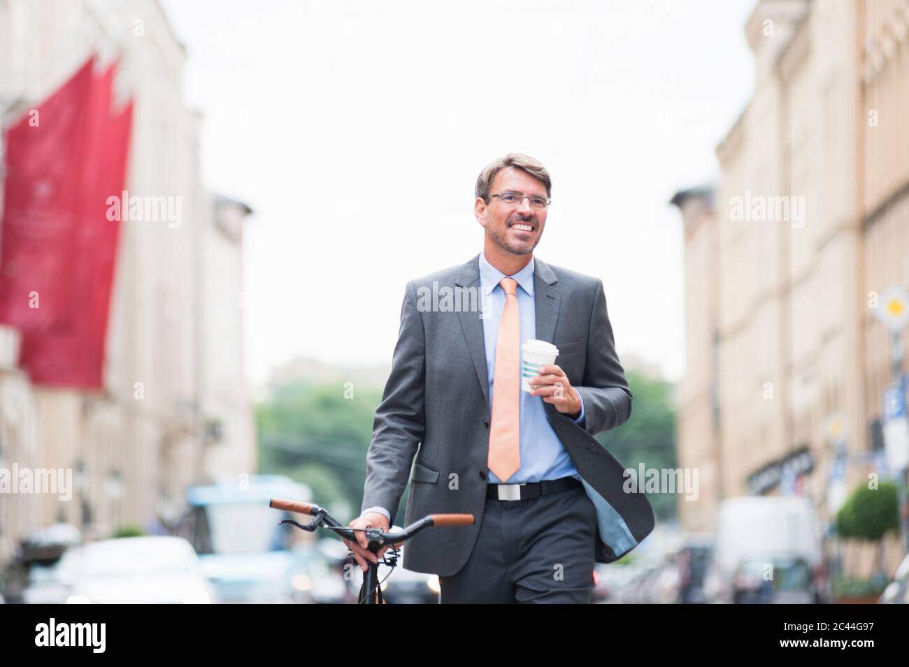 Smiling businessman holding coffee looking away while walking with bicycle in city Stock Photo