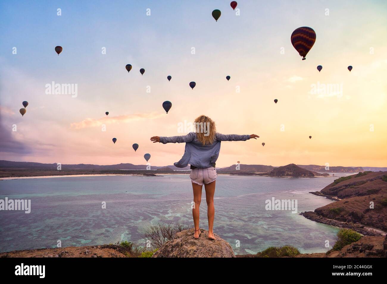 Indonesia, West Nusa Tenggara, Hot air balloons flying over lone woman standing on rocky shore with raised arms Stock Photo