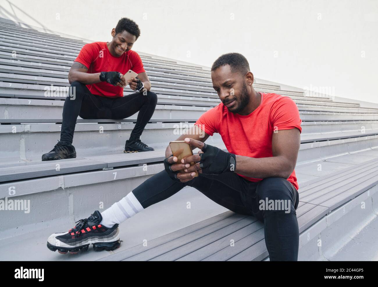 Two sportsmen sitting on stairs in a stadium using smartphones Stock Photo