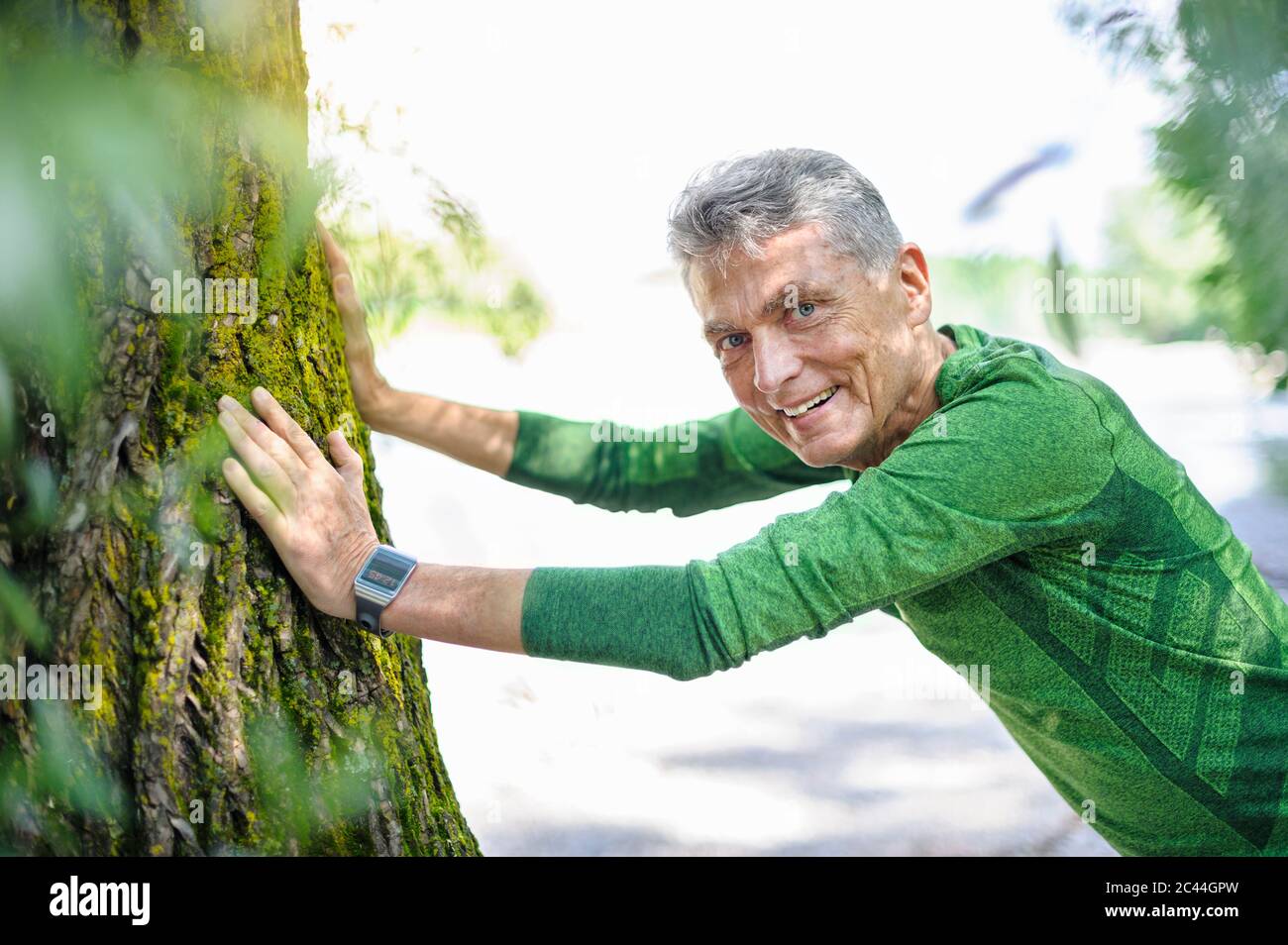 Portrait of smiling retired elderly man exercising while leaning on tree trunk at park Stock Photo