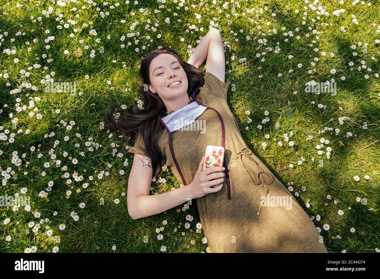 Woman with let down face mask enjoying her free time while lying on grass with daisies Stock Photo