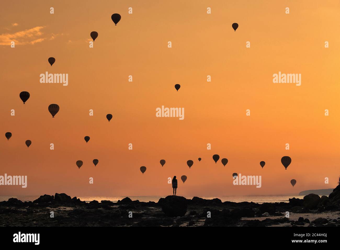 Indonesia, West Nusa Tenggara, Silhouettes of hot air balloons flying over lone woman standing on rocky shore at moody dusk Stock Photo
