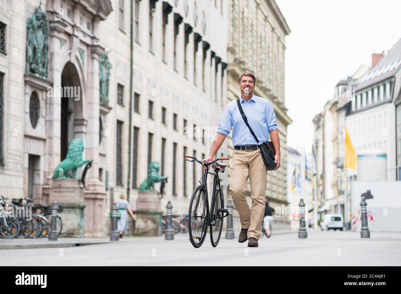 Smiling businessman carrying messenger bag walking with bicycle on street in city Stock Photo