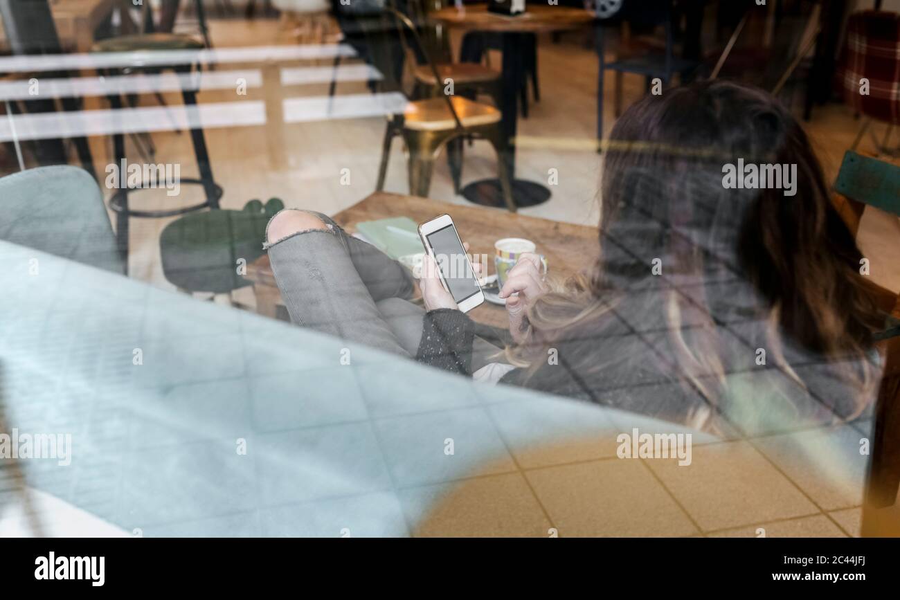 Woman using smartphone in coffee shop, seen through windowpane Stock Photo