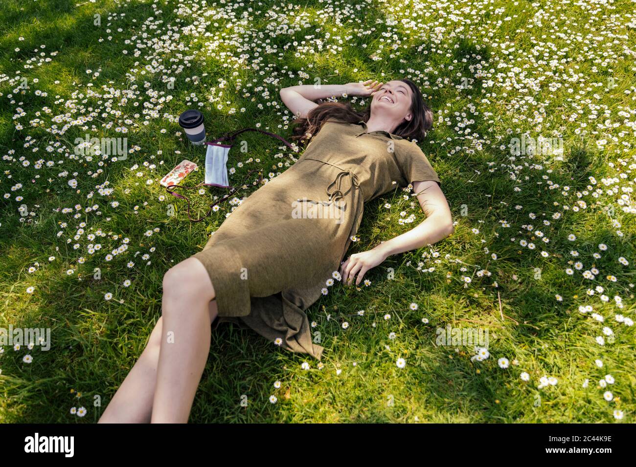 Happy woman enjoying her free time while lying on grass with daisies next to face mask Stock Photo