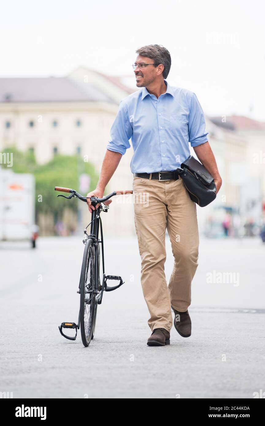 Smiling businessman looking away while walking with bicycle on street in city Stock Photo