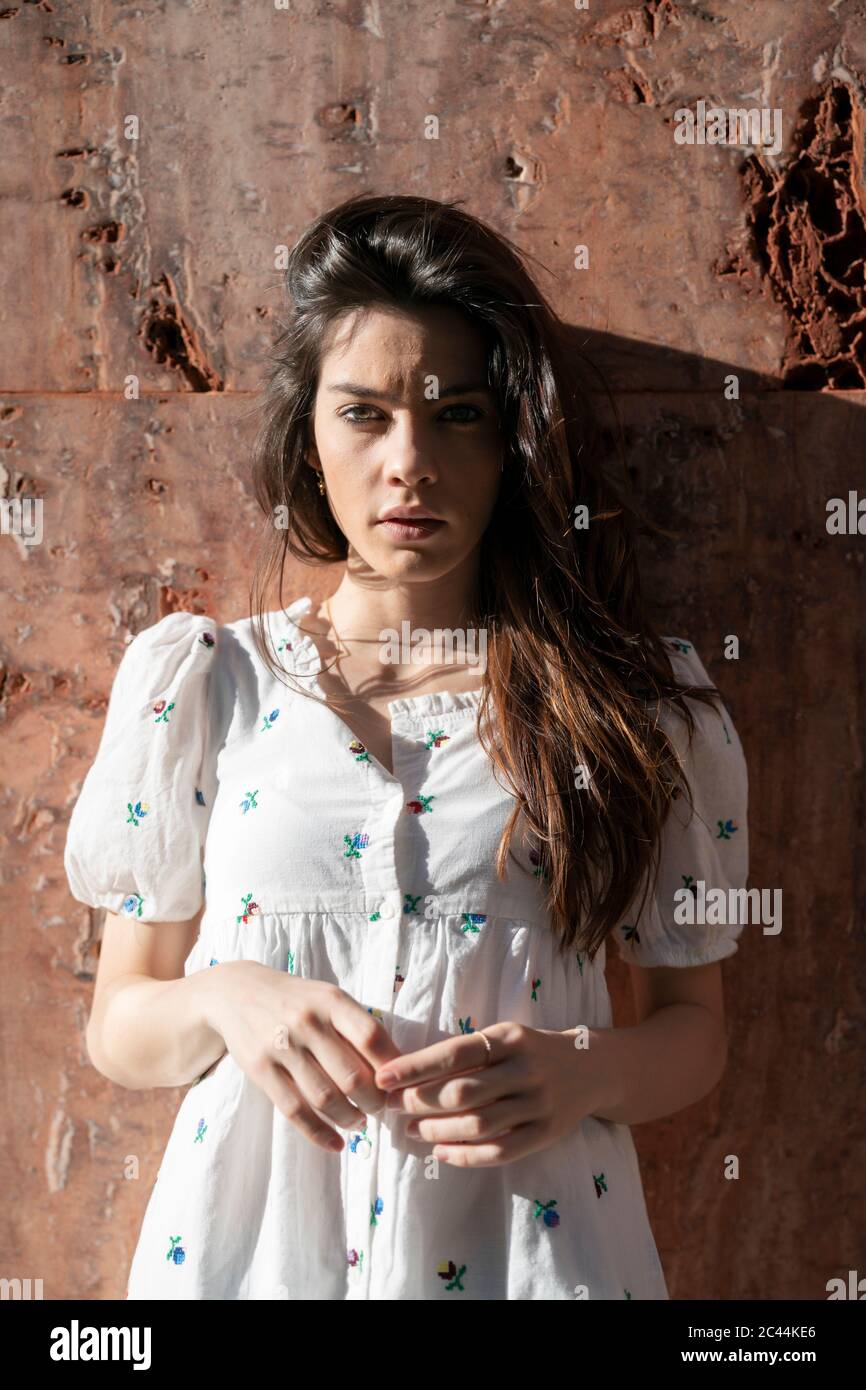 Young woman in white mini dress standing against weathered wall during sunny day Stock Photo