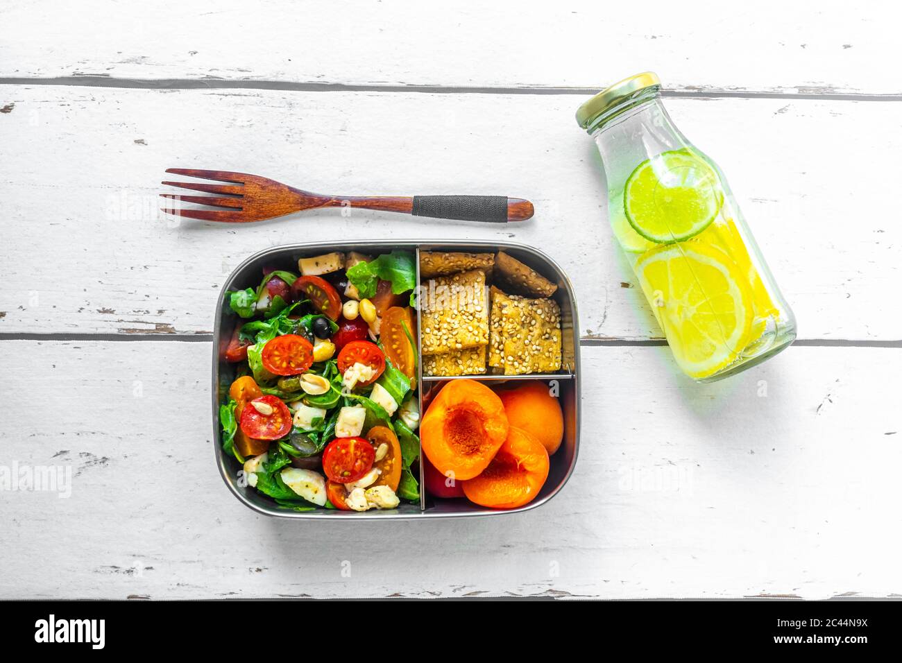 Lunch box with rocket salad with colored tomatoes, mozzarella and nuts, crispbread and apricots and bottle of infused water with lemons and slices of lime Stock Photo