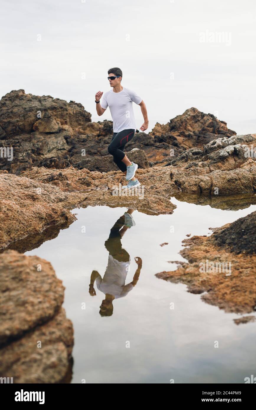 Full length of male trail runner running on rocks with his reflection over water at coast, Ferrol, Galicia, Spain Stock Photo