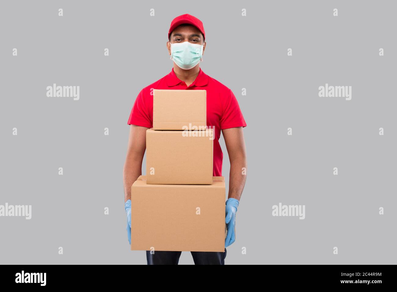 Delivery Man Holding Carton Boxes WEaring Medical Mask and Gloves Isolated. Indian Delivery Boy Smiling with Boxes in Hands Stock Photo