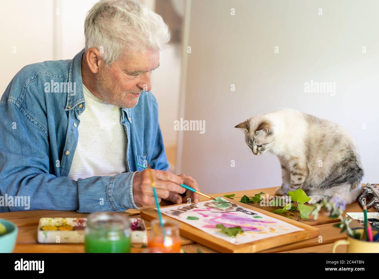 Senior man painting on paper while cat sitting on table at home Stock Photo