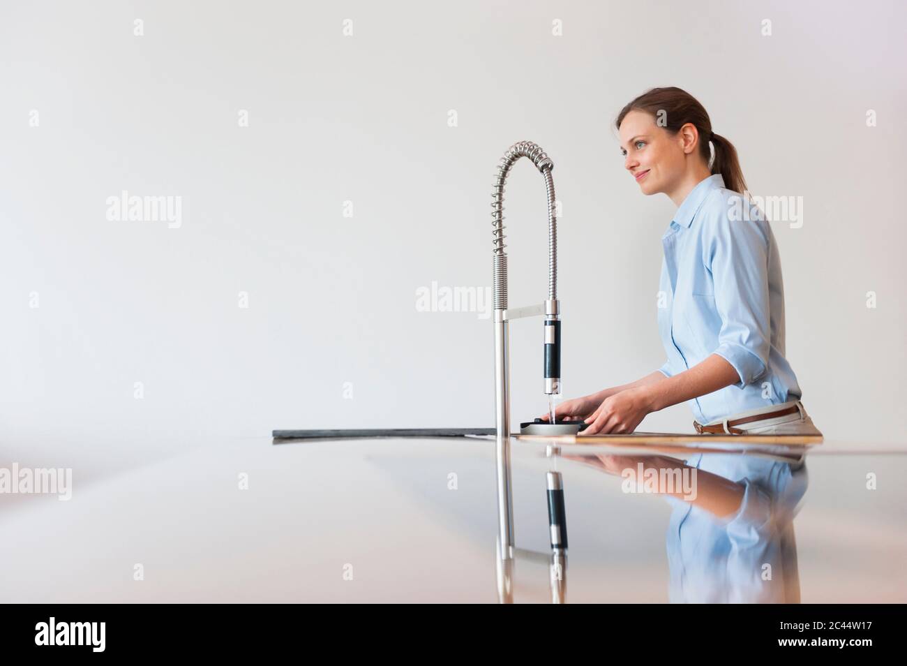 Smiling woman filling water in container through modern kitchen faucet against wall Stock Photo