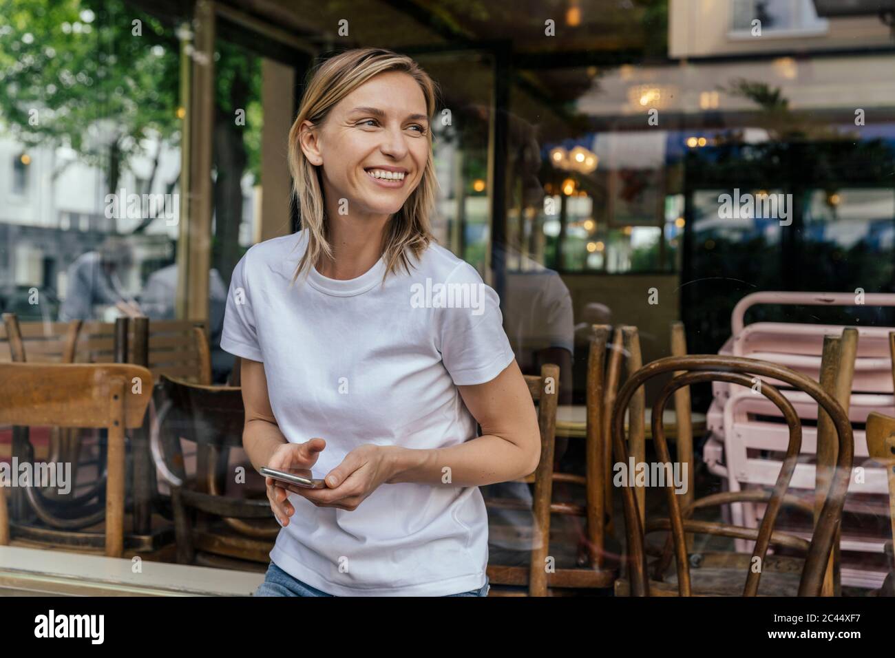 Portrait of smiling woman with smartphone standing in front of a closed coffee shop looking at distance Stock Photo