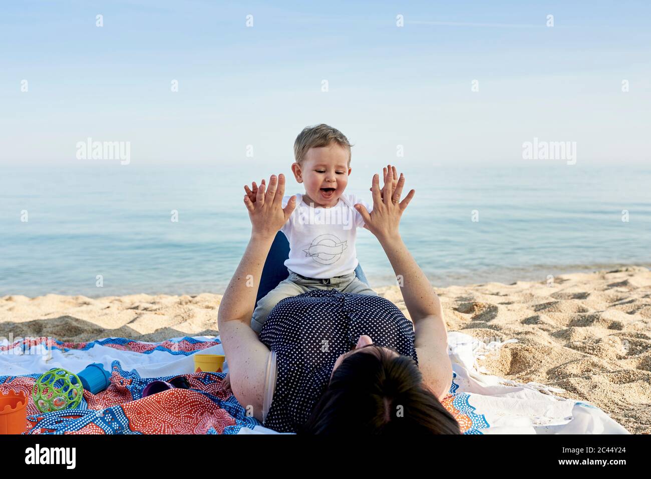 Mother playing clapping game with son while lying on blanket at beach against sea Stock Photo