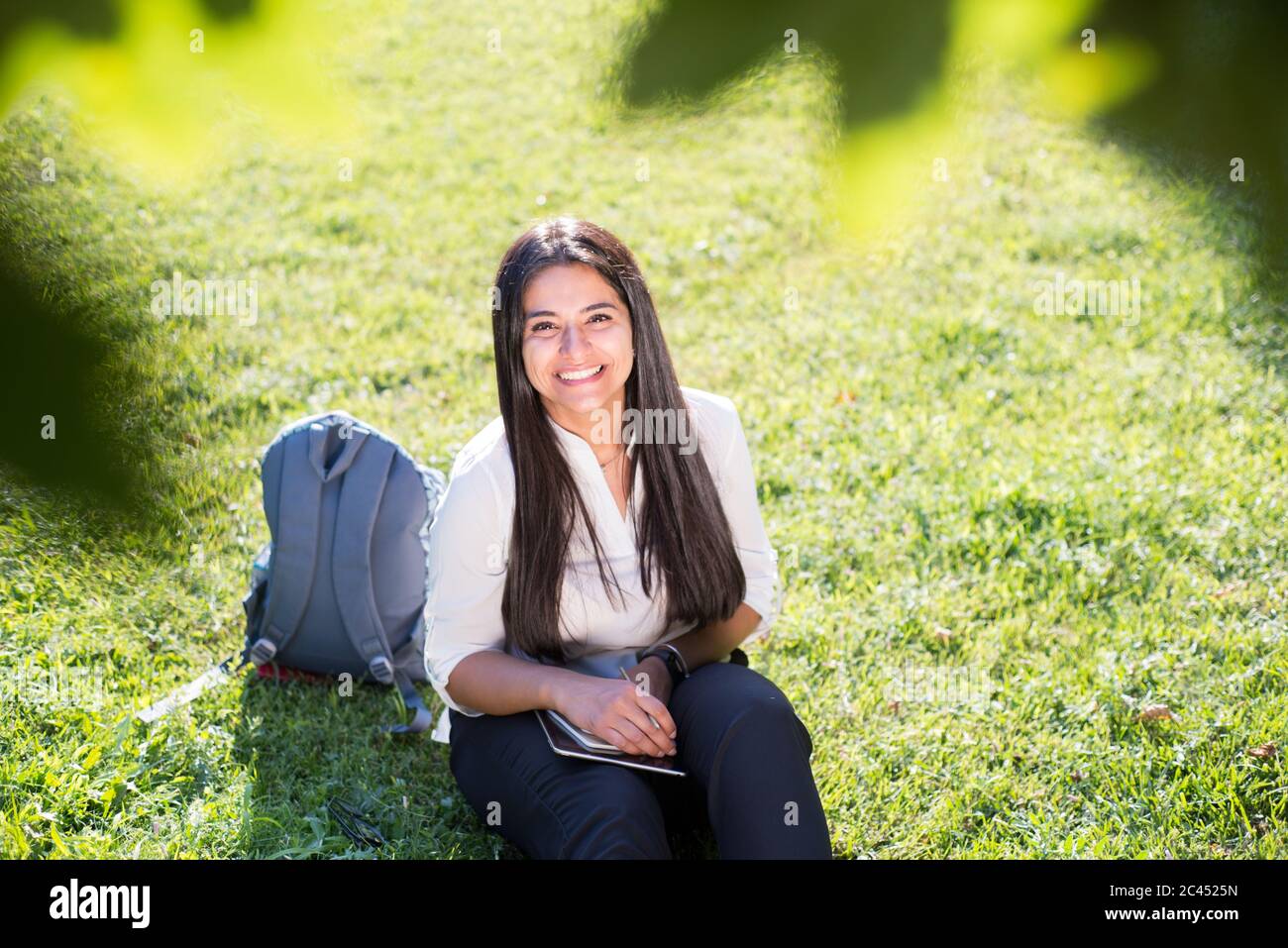 Indian girl, student, smiles, sits on the green grass, with a backpack and notepad, writes Stock Photo