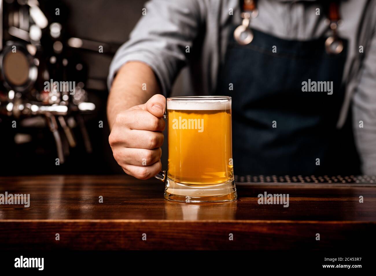 Barman in apron serves beer in glass mug on wooden bar counter Stock Photo
