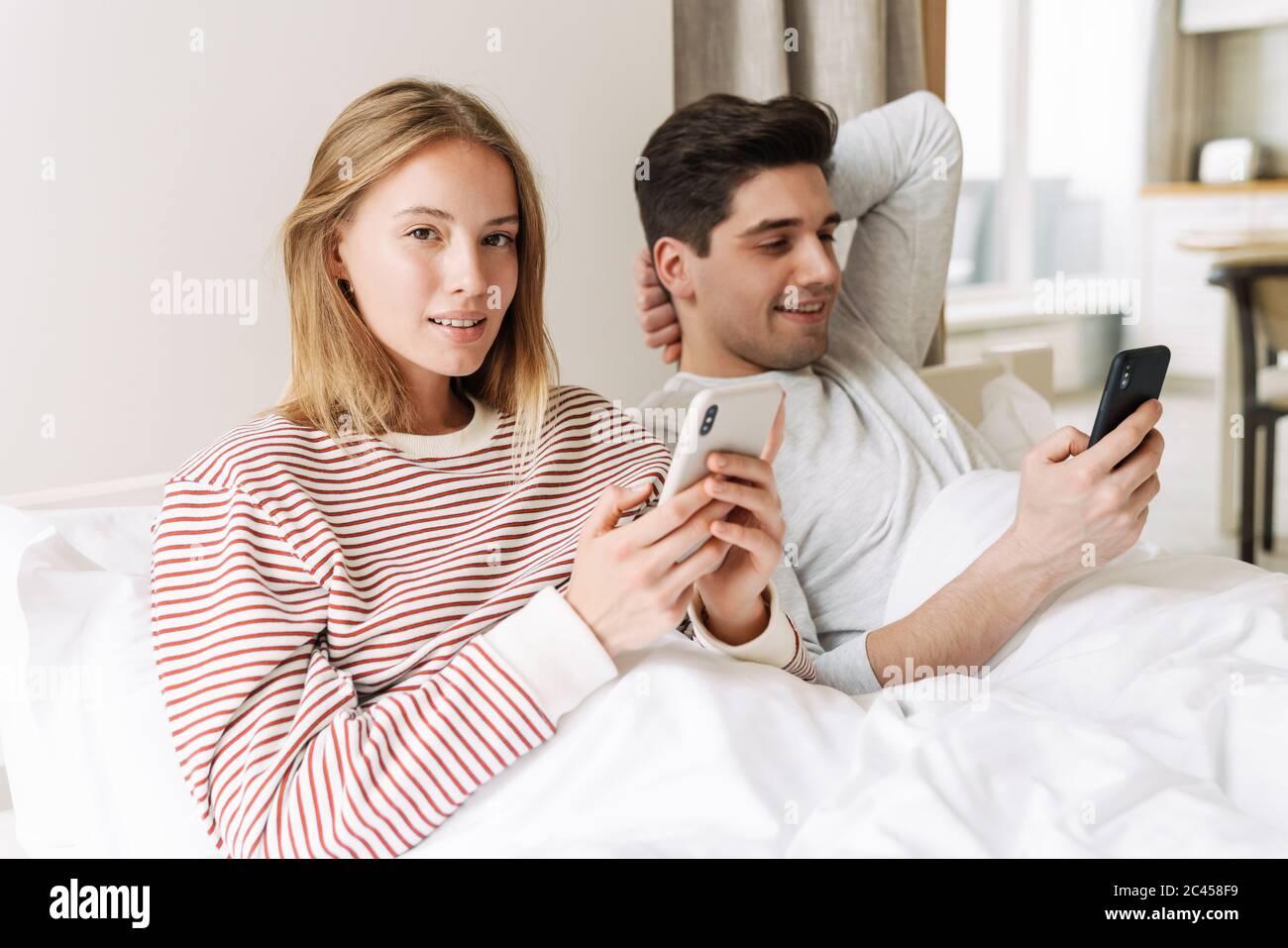 Portrait of beautiful happy couple smiling and using smartphones while lying in bed at home Stock Photo