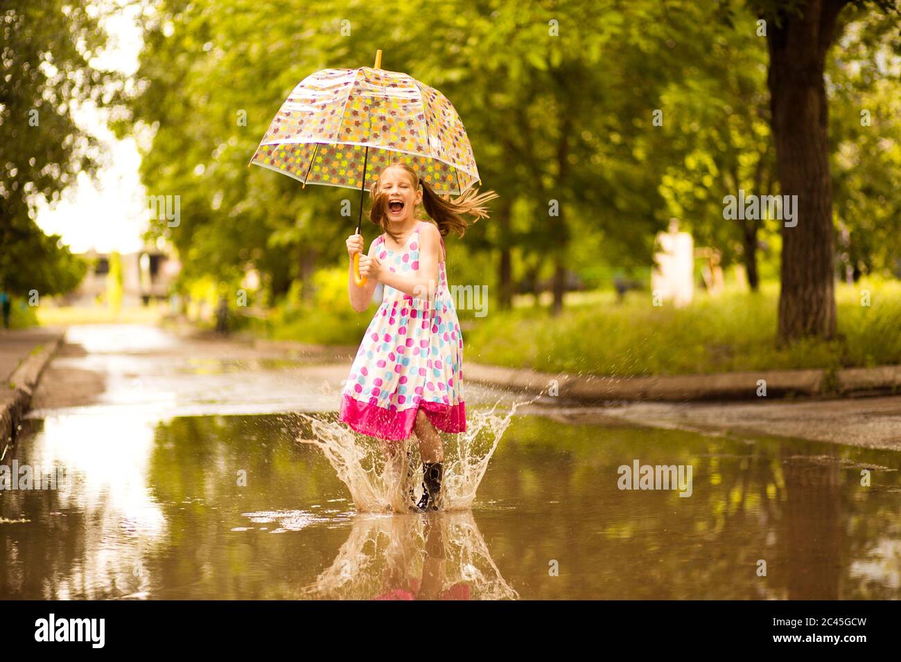 Happy funny kid girl with umbrella jumping on puddles in rubber boots ...