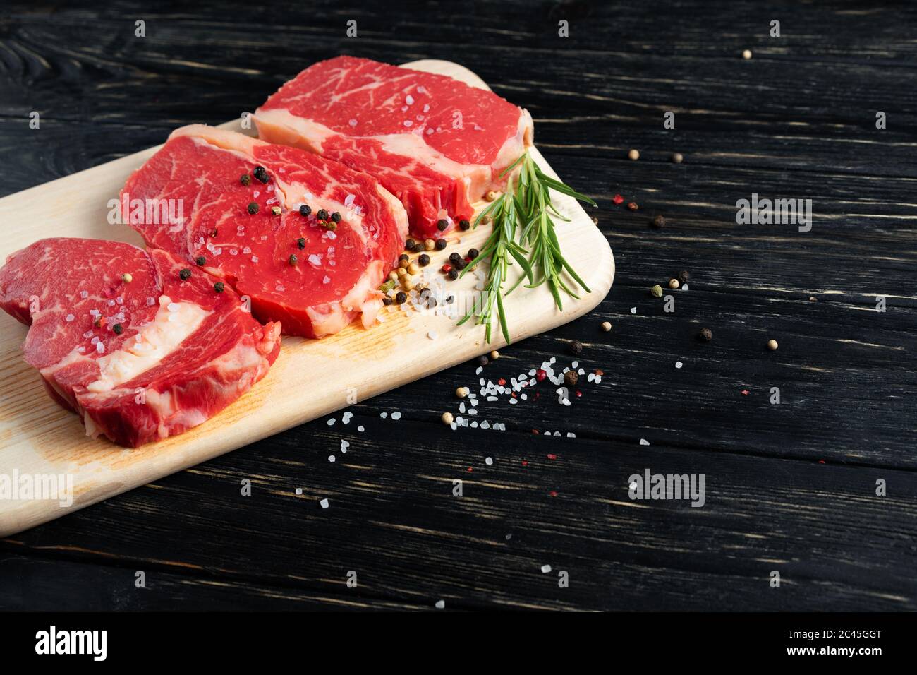 Three pieces of juicy raw beef with rosemary on a cutting board on a black wooden table background. Meat for barbecue or grill sprinkled with pepper and salt seasoning Stock Photo