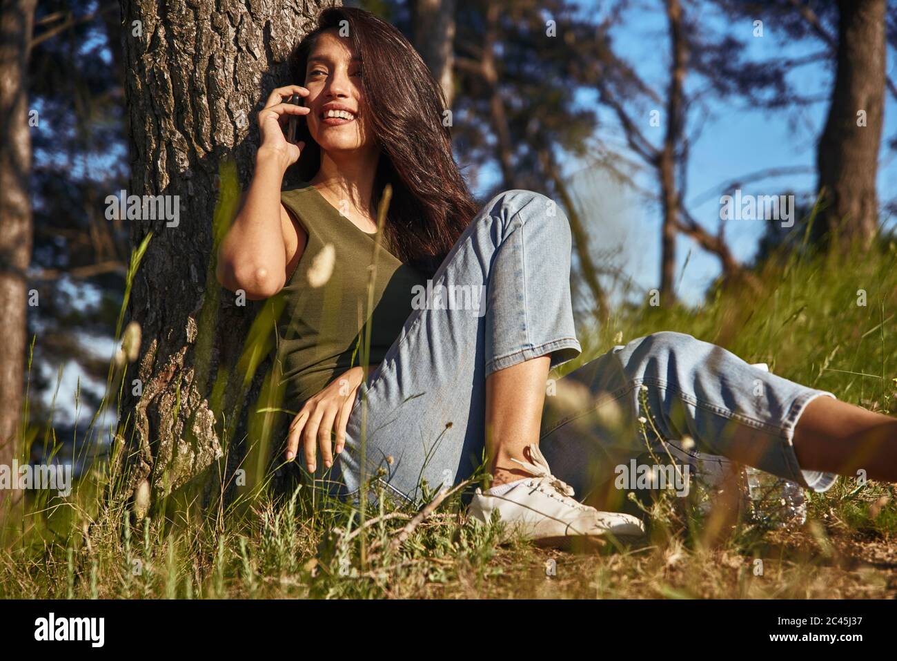 Portrait of young woman with long brown hair sitting under a tree in a forest, talking on mobile phone. Stock Photo