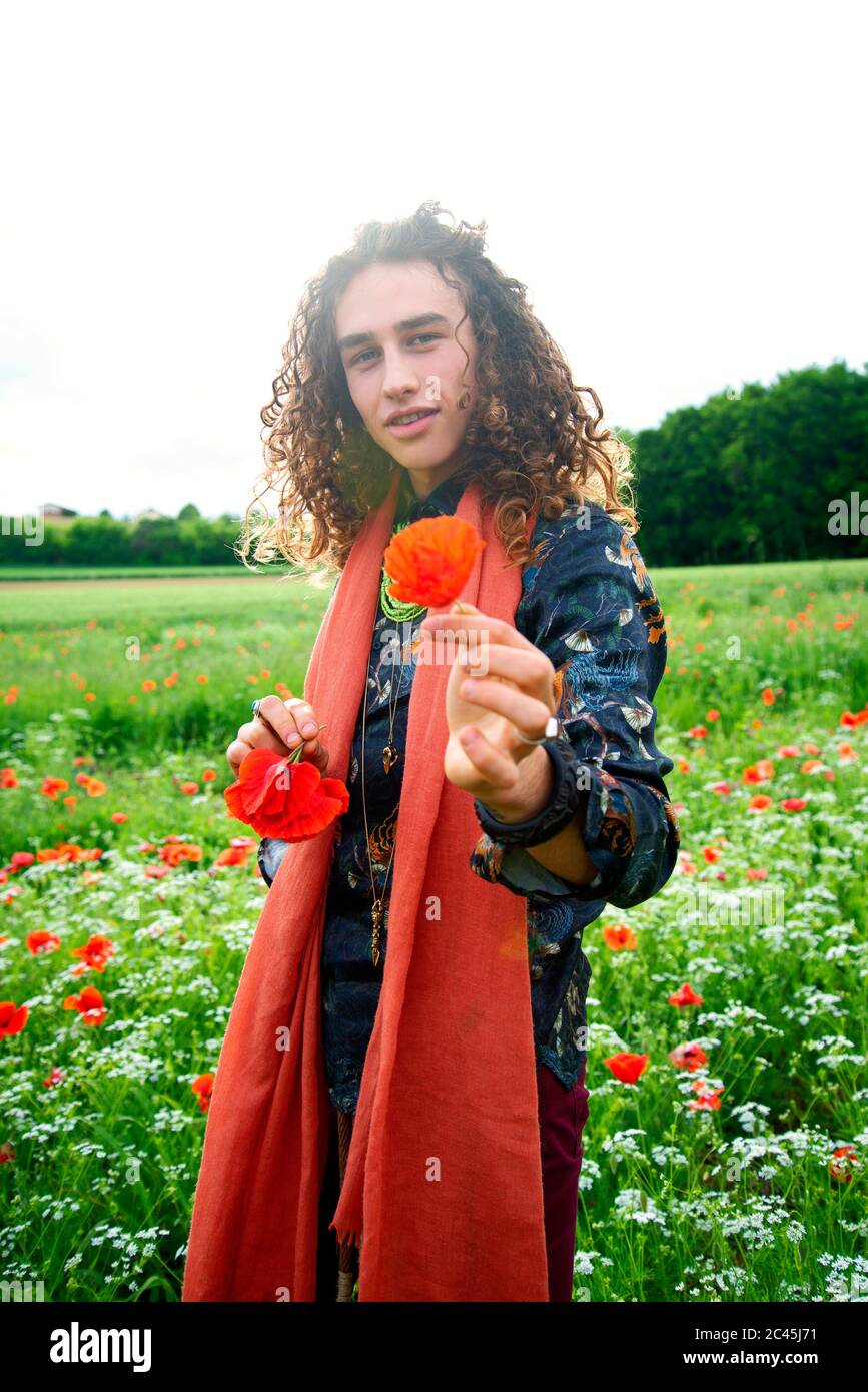 Portrait of young man with long brown curly hair standing in a poppy meadow. Stock Photo
