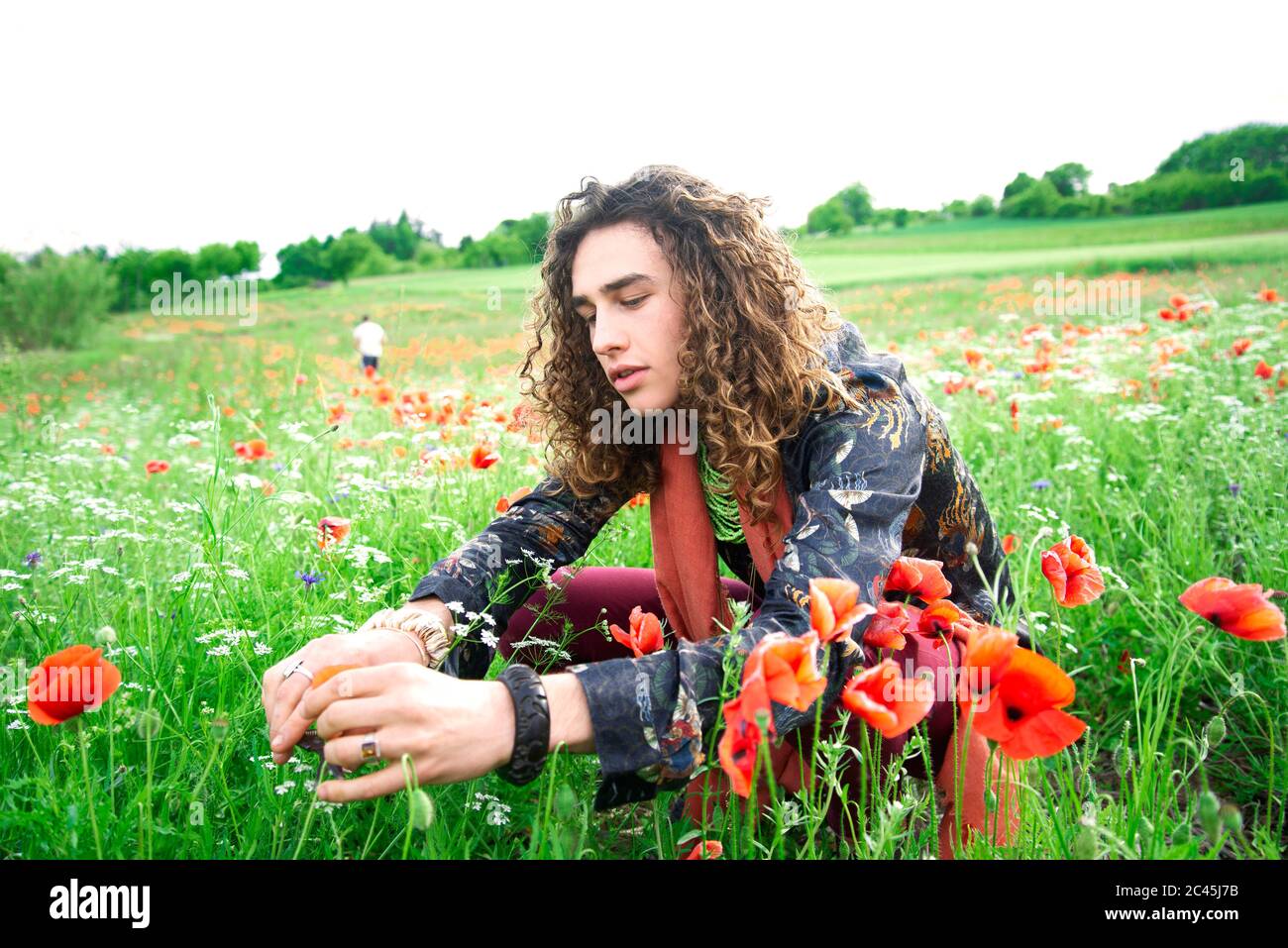 Portrait of young man with long brown curly hair sitting in a poppy meadow Stock Photo