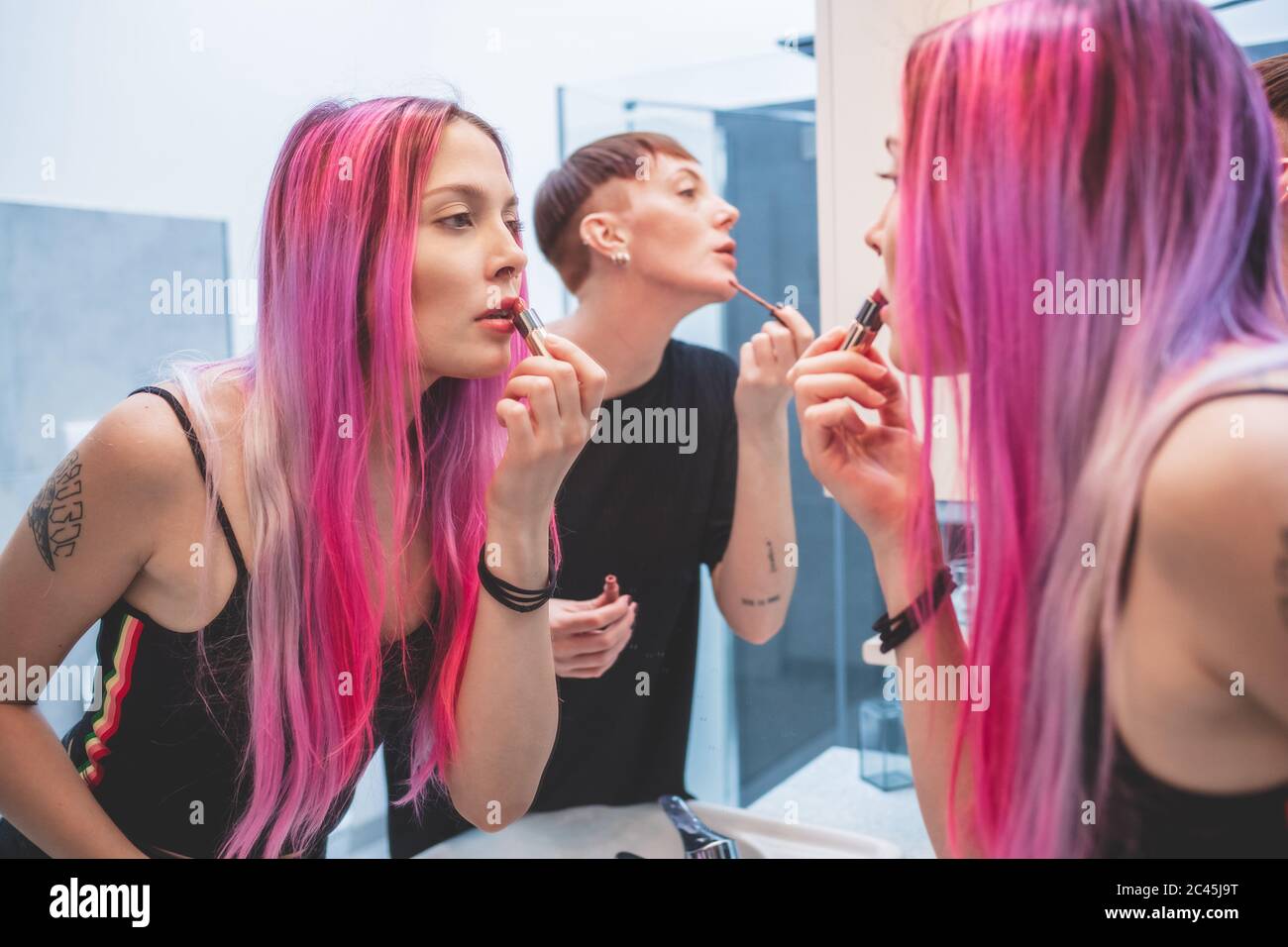 Young woman with long pink hair and woman with short red hair standing in  front of mirror, applying lipstick Stock Photo - Alamy