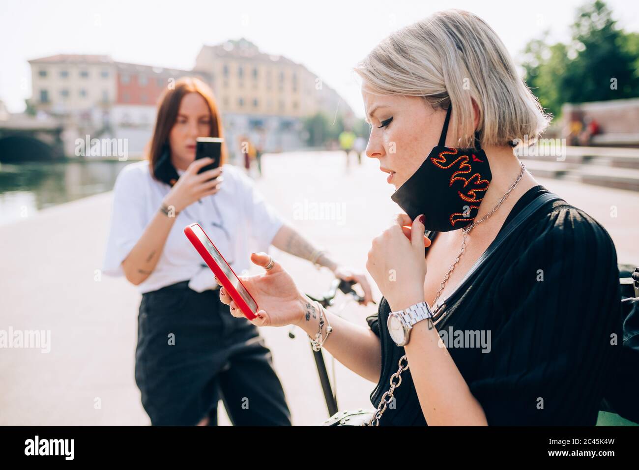 Two young women wearing face masks during Corona virus, standing outdoors, using mobile phone. Stock Photo
