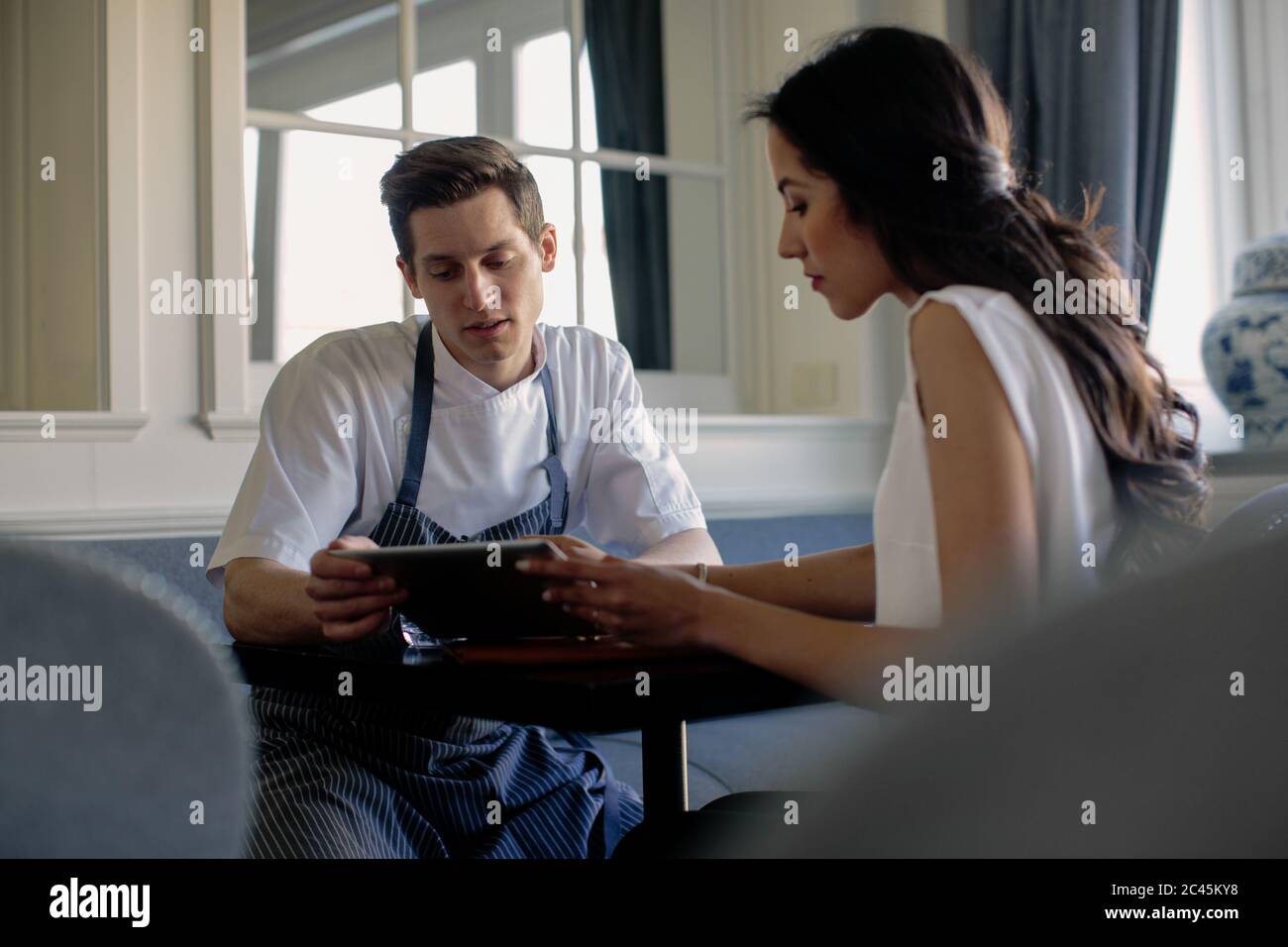 Chef wearing blue apron and woman sitting at a table, looking at digital tablet. Stock Photo