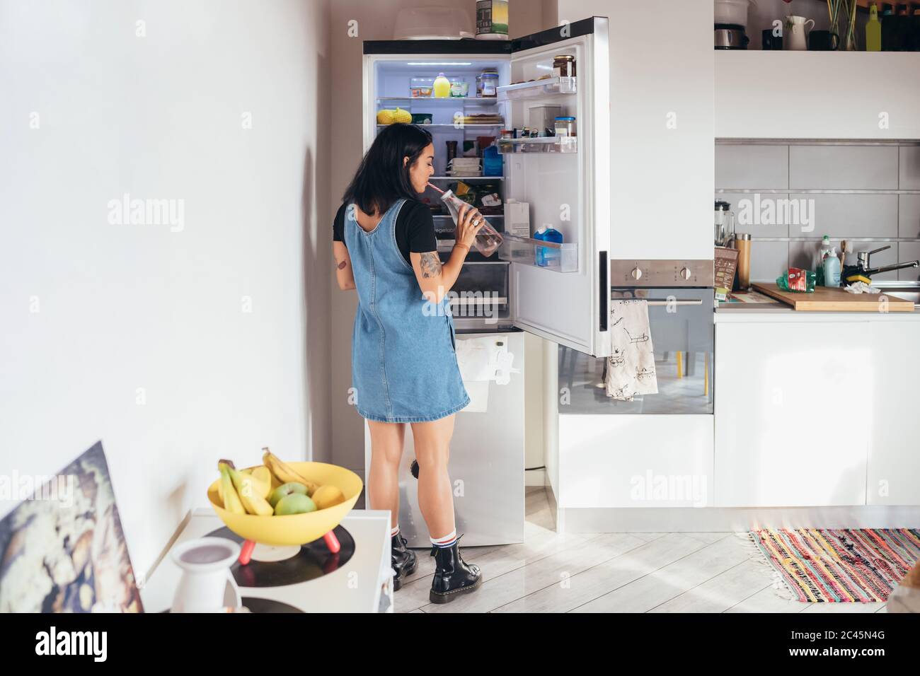 Rear view of tattooed woman with long brown hair wearing denim dress standing in front of open fridge, drinking from bottle. Stock Photo