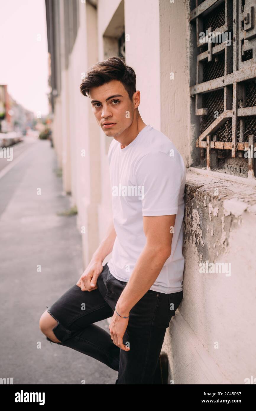 Portrait of young man with dark brown hair, wearing white T-shirt and  ripped black jeans, leaning against wall Stock Photo - Alamy