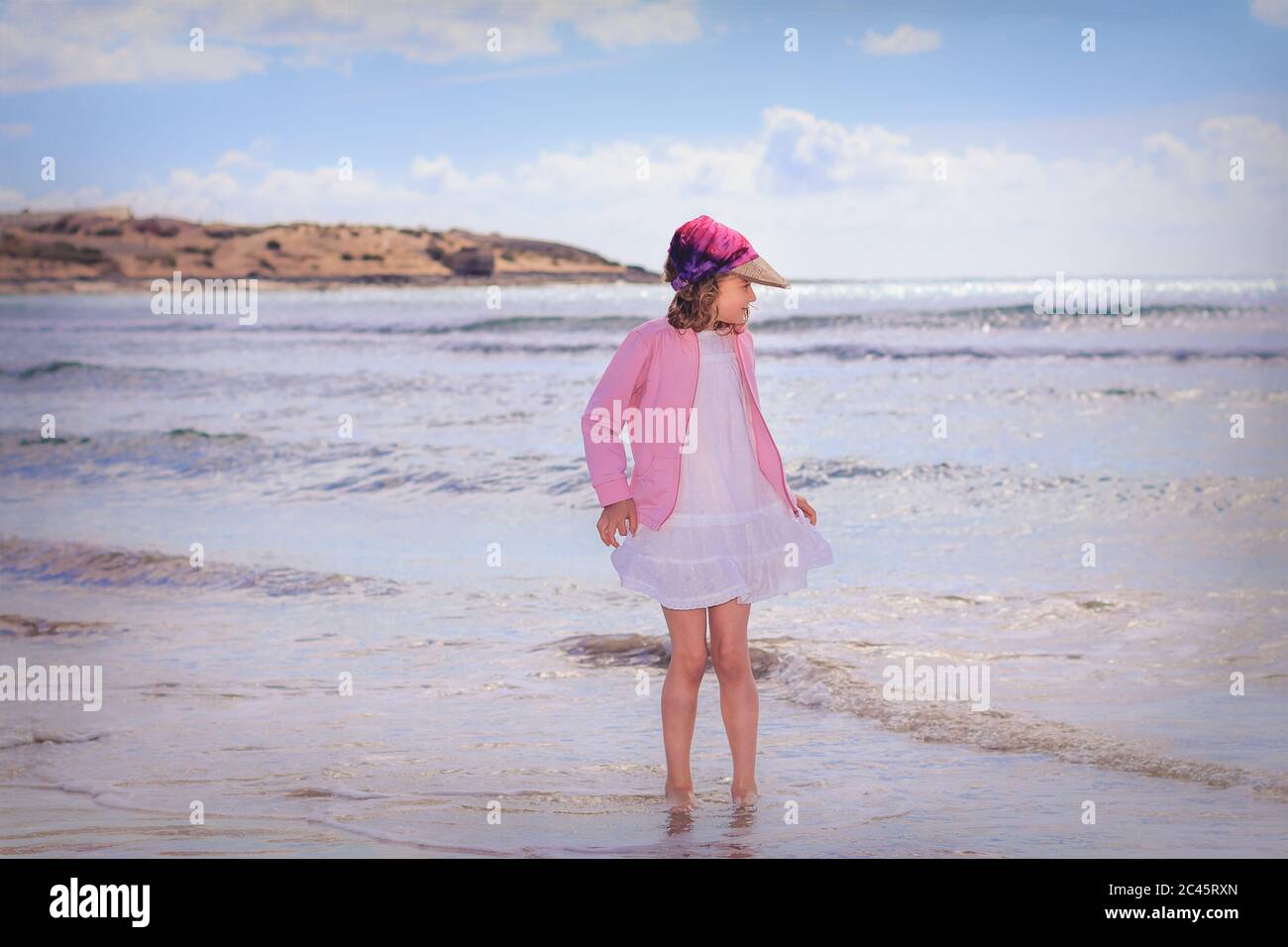 Seascape image of young girl wearing white dress standing with feet in water Stock Photo