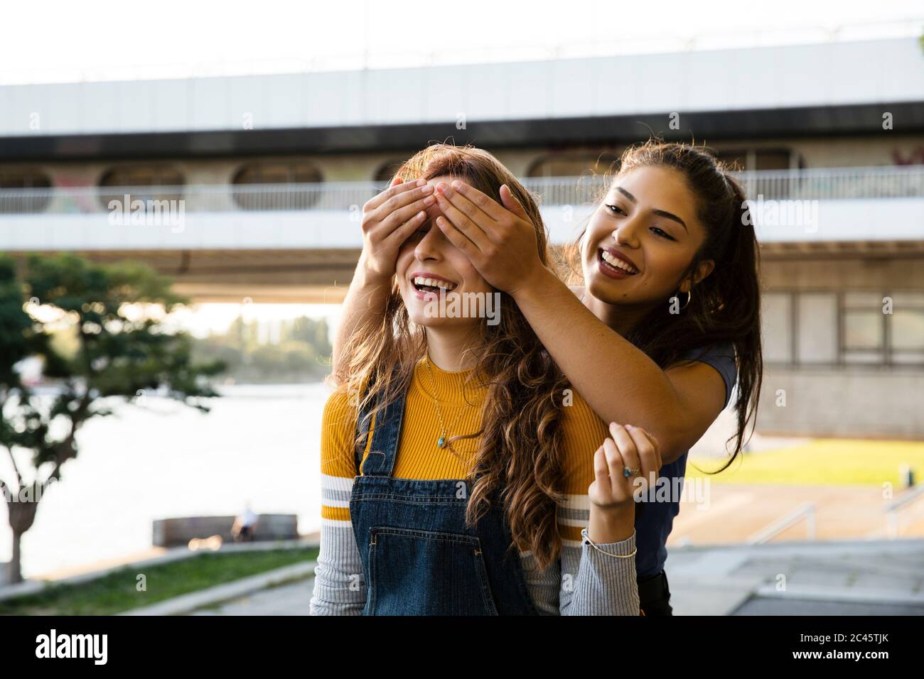 Two young women with long brown hair standing on riverbank, smiling and ...
