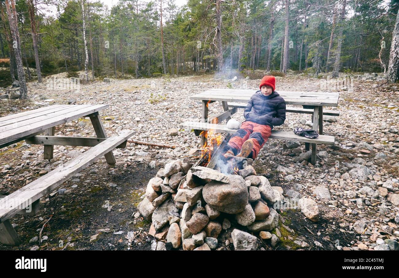 Boy sitting on picnic bench next to campfire in a forest in Vasterbottens Lan, Sweden. Stock Photo