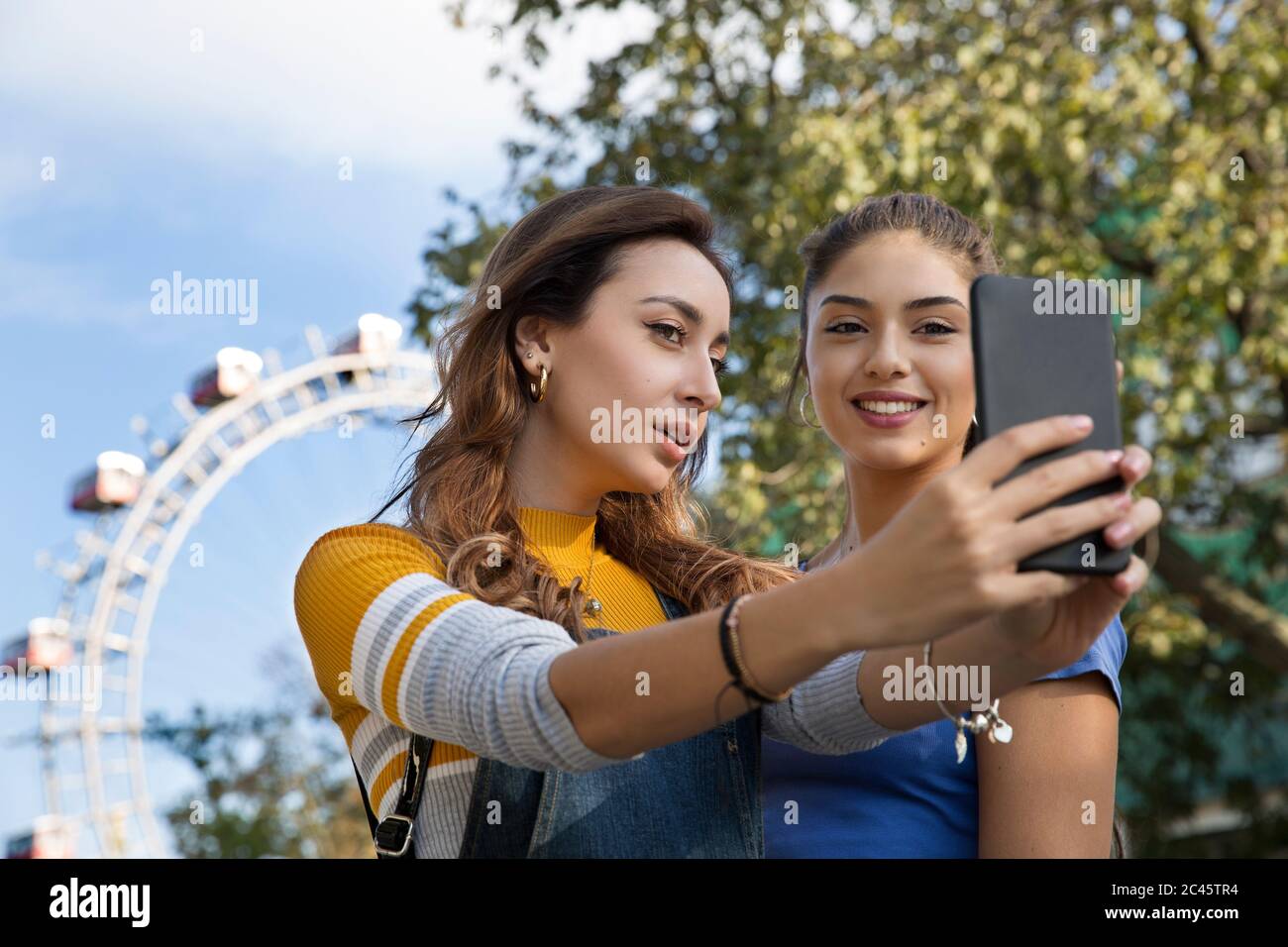 Two young women with long brown hair standing in a park near a Ferris wheel, taking selfie with mobile phone. Stock Photo