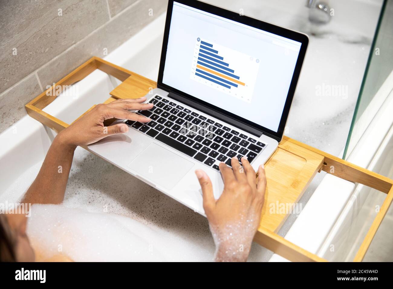 Woman sitting in bathtub, having foam bath and working on laptop during Coronavirus crisis. Stock Photo