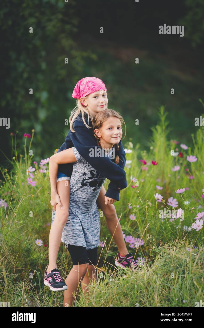 Young tween girl carrying friend piggyback in wild flower meadow Stock Photo