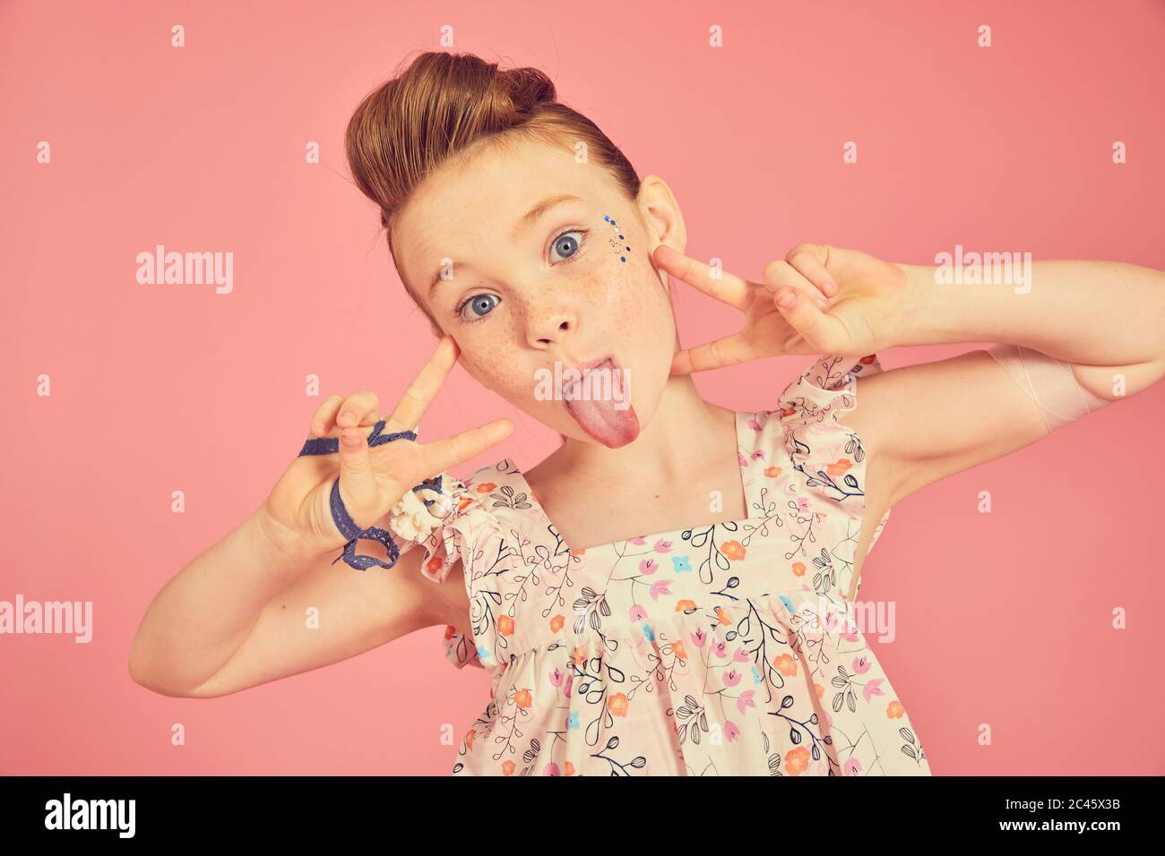 Portrait of brunette girl wearing frilly dress with floral pattern on pink background, sticking out tongue at camera. Stock Photo