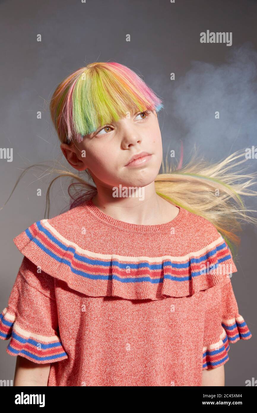 Portrait of girl with long blond hair and dyed fringe wearing pink frilly top, looking up, on grey background. Stock Photo