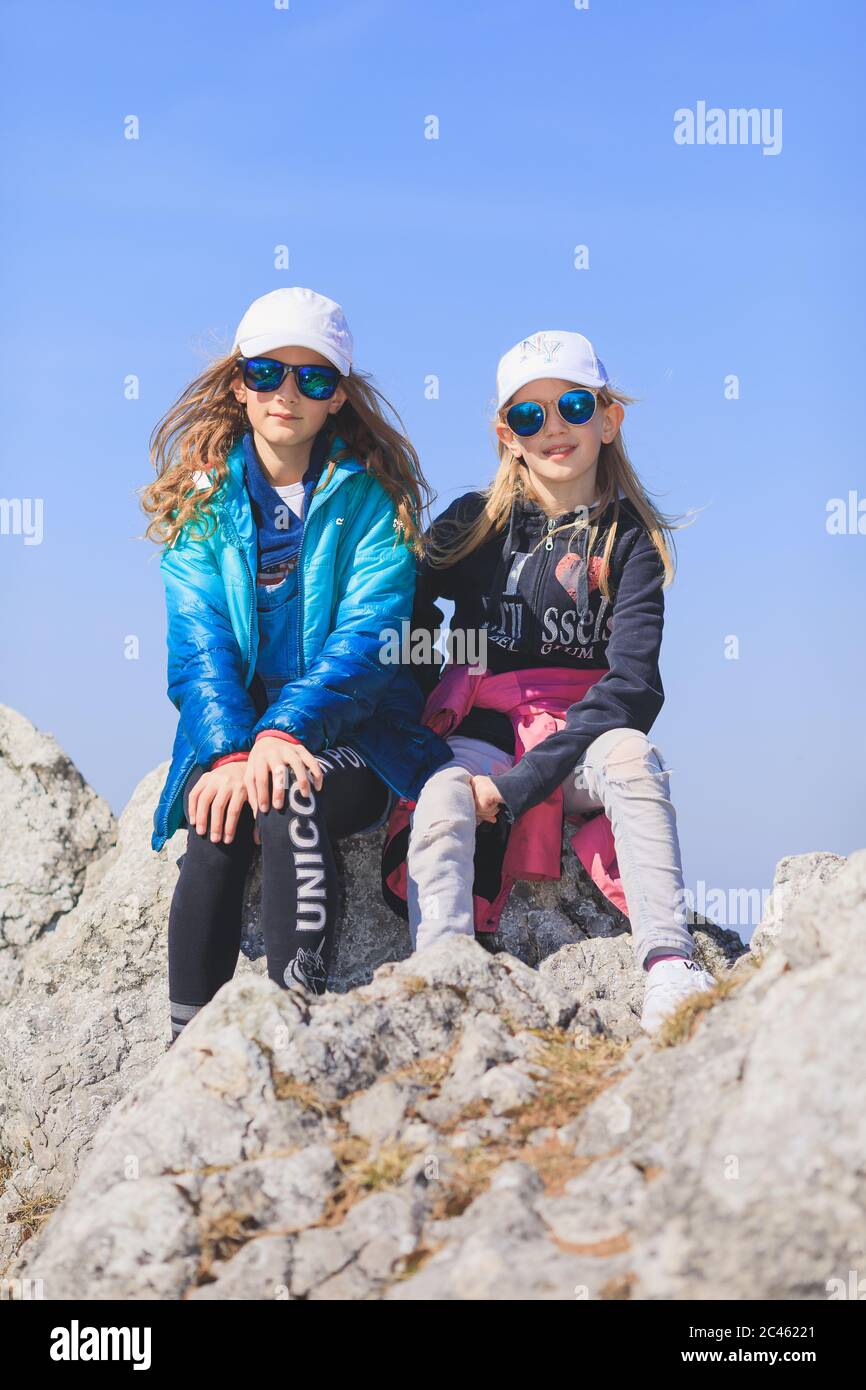 Two young girls wearing sun caps and sunglasses sitting on top of mountain Stock Photo