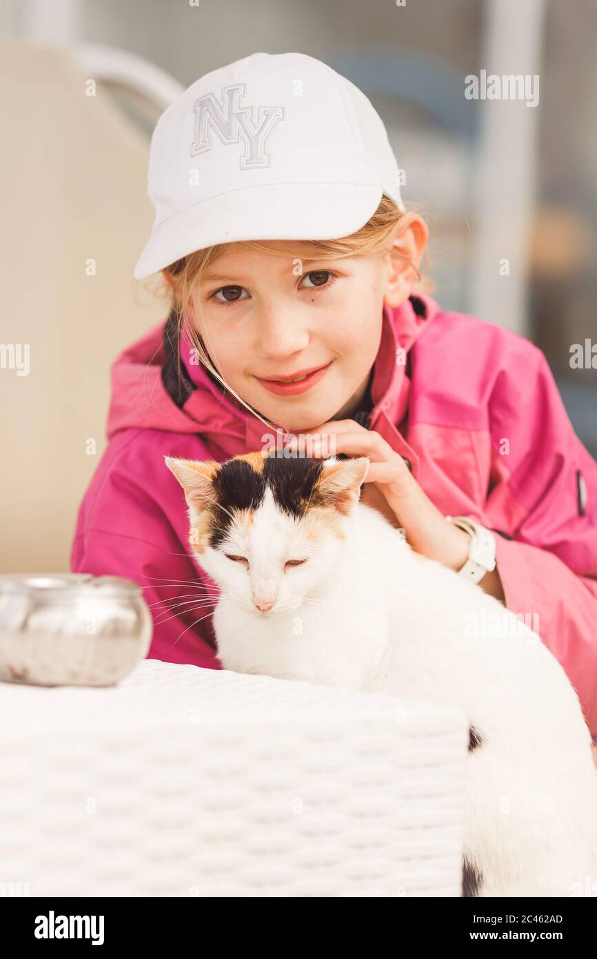 Young blond girl, primary school age, wearing sun cap, caressing cat lying on sunbed Stock Photo