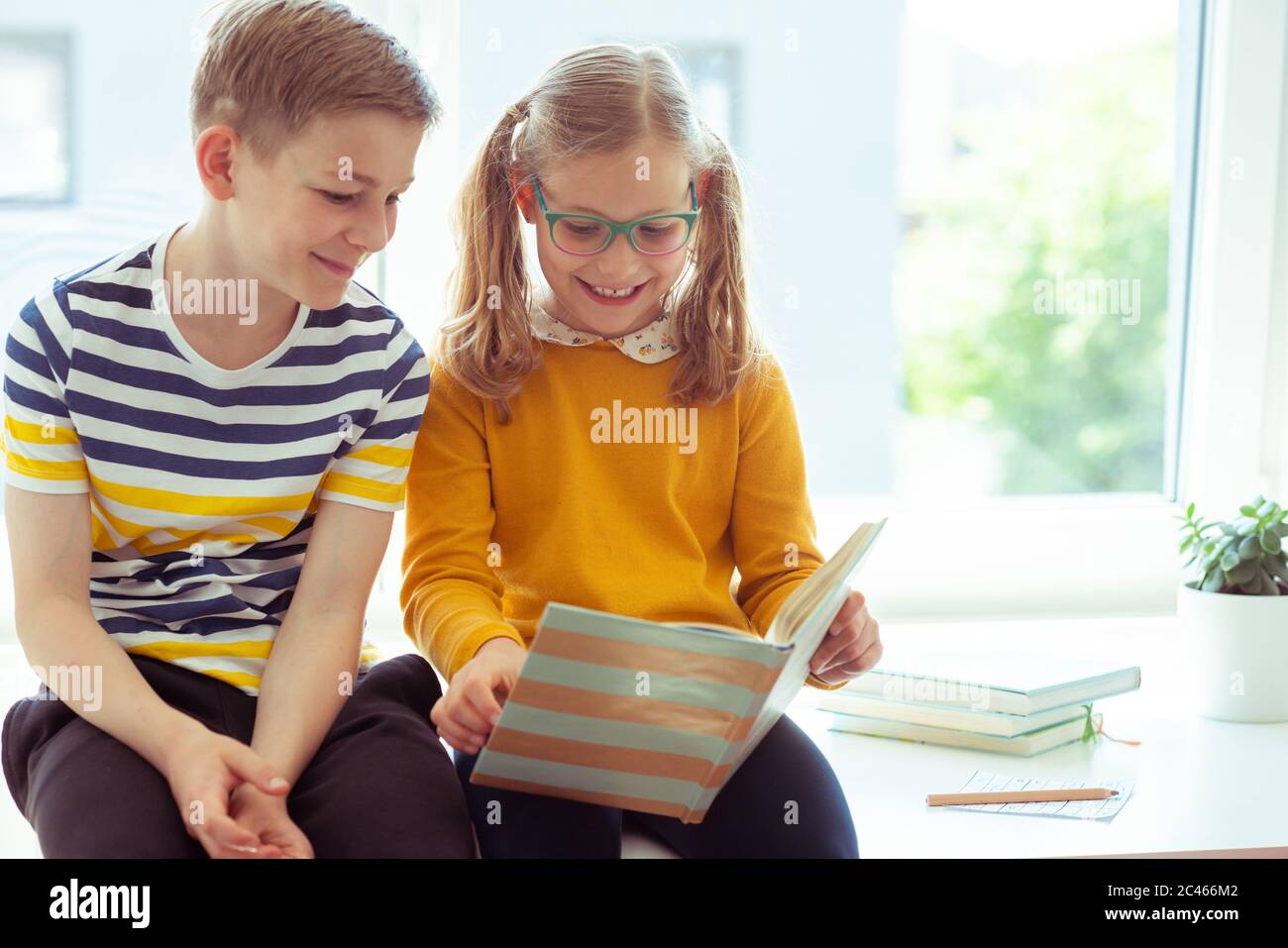 Two happy teenagers children sitting on desk and reading a funny book ...