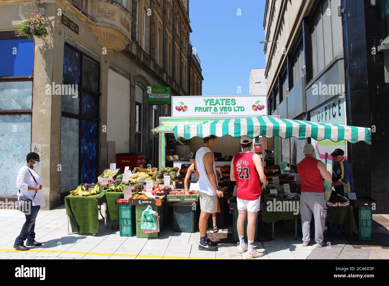 A photograph of a fruit and veg trader on Cardiff Queen Street.  Customers following social distancing measures queuing up.  Farmer's market. Stock Photo