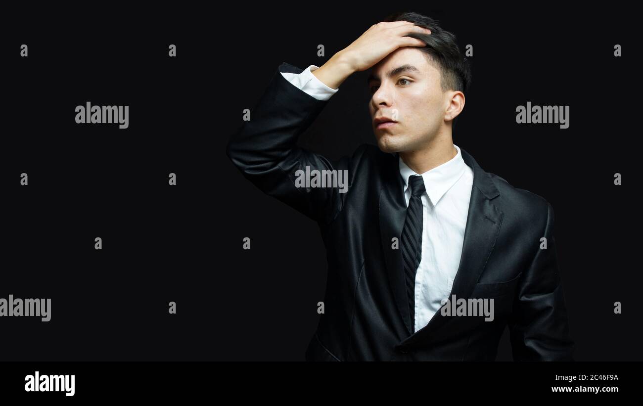 Young latin man in black suit, white shirt, black tie in worried attitude with his hand on his head on black background Stock Photo