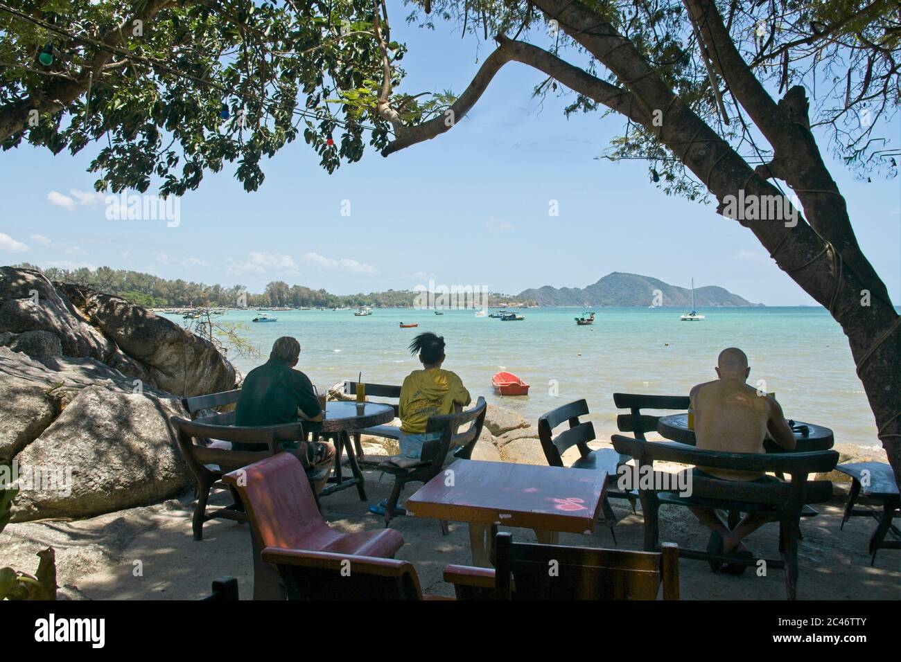 Secluded bar & boats on sandy beach people seated al-fresco framed by tree branch leaves and trunk looking out to sea blue sky turquoise sea Landscape Stock Photo