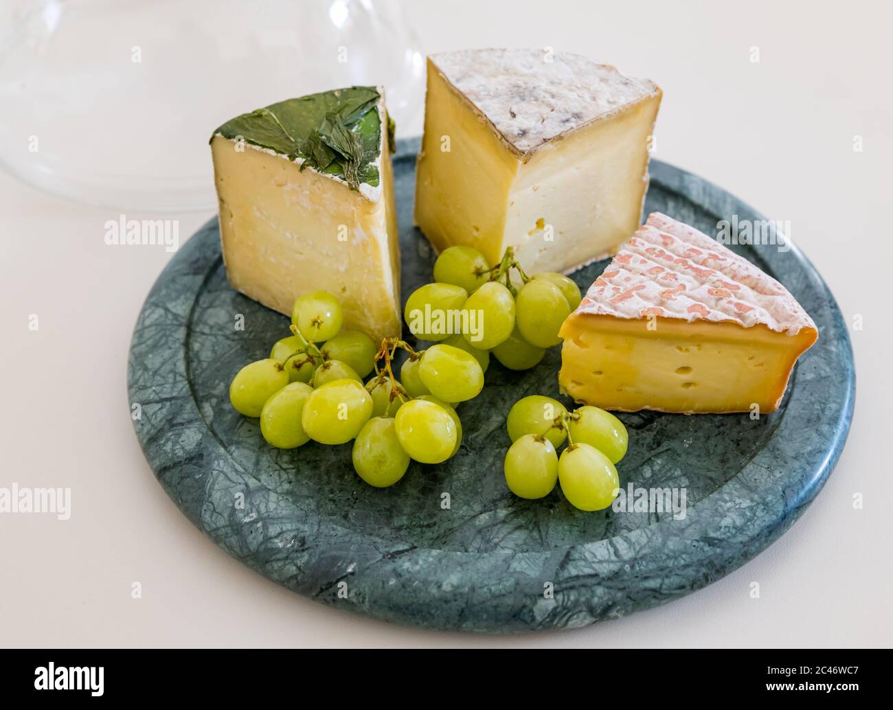 Cheeseboard with Garlic Yarg, Gorwydd Caerphilly and Irish Gubbeen cheeses with green grapes on white kitchen counter Stock Photo